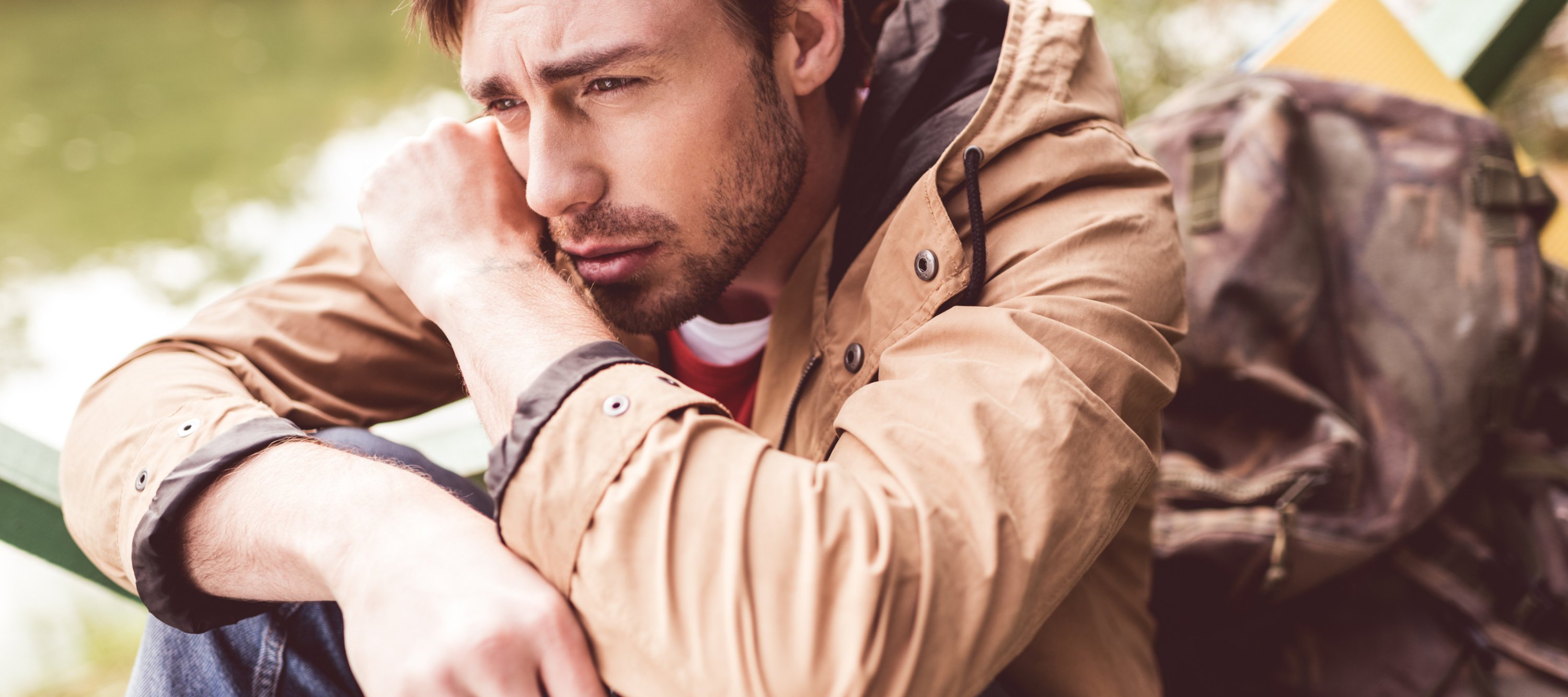 Close-up portrait of young bearded thoughtful man traveler with backpack sitting on pier near river