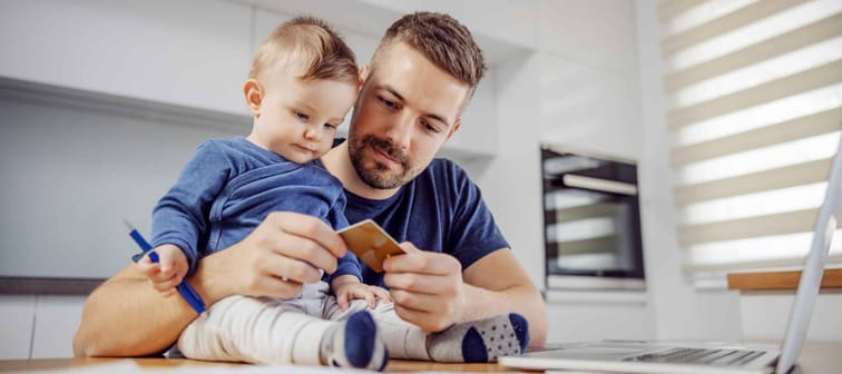 Young attractive bearded father sitting at dinning table with his beloved adorable son and paying bills online. His boy holding pen and trying to help him.