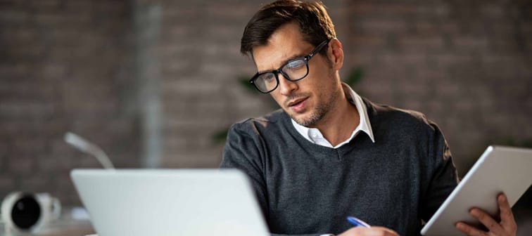 Multi-tasking businessman working in the office. He is using touchpad while reading an e-mail on laptop and taking notes on the paper.
