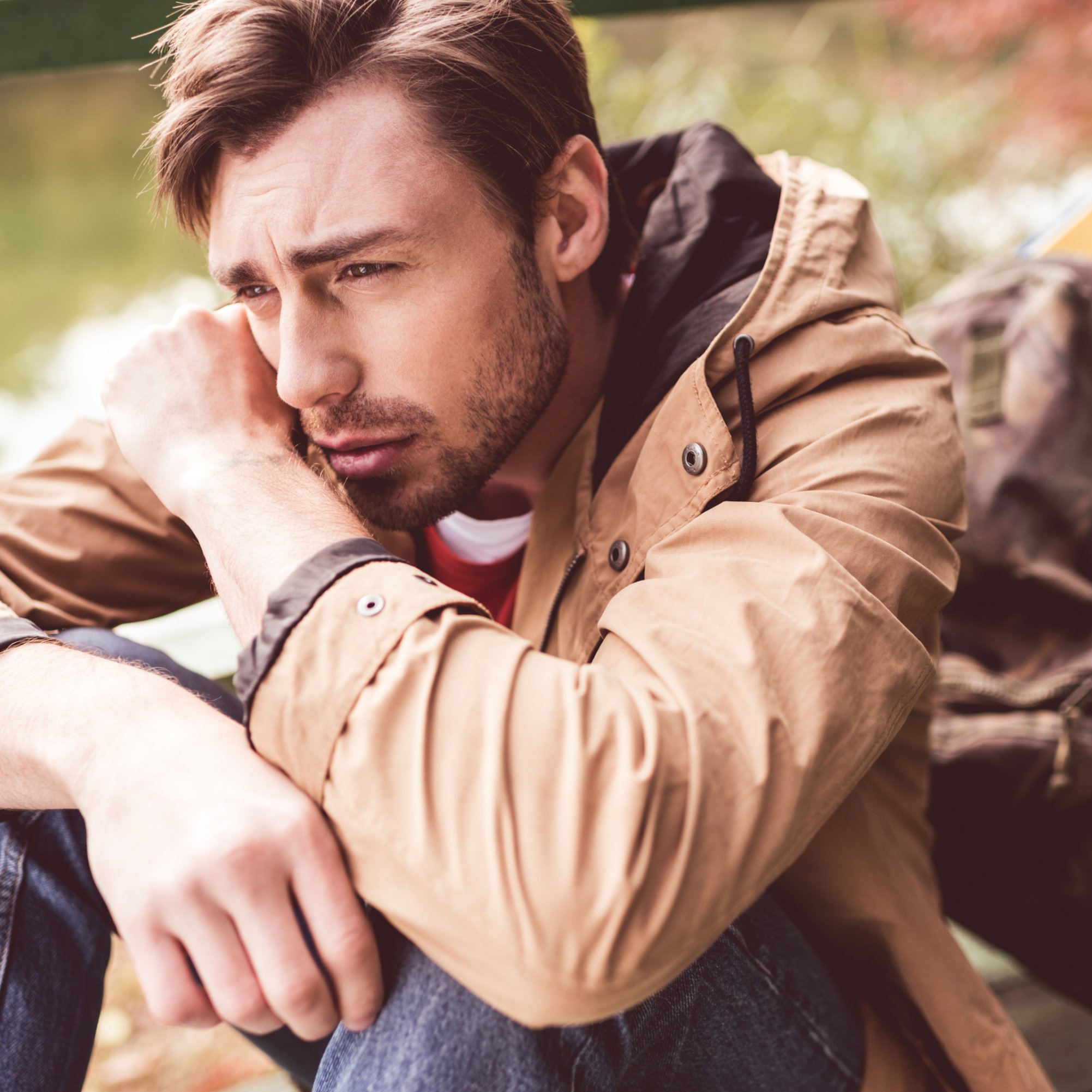Close-up portrait of young bearded thoughtful man traveler with backpack sitting on pier near river