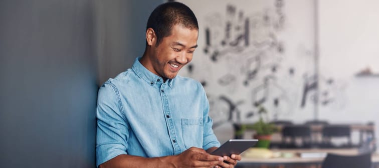 Casually dressed young Asian designer smiling and using a digital tablet while leaning against a gray wall in a modern office